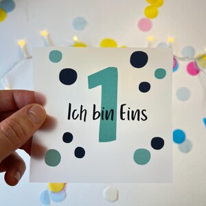 Könnte beinhalten: Eine weiße Karte mit der Zahl 1 in Türkis und den Worten "Ich bin Eins" in Schwarz. Die Karte ist mit türkisfarbenen und marineblauen Punkten verziert. Bunte Konfetti und Lichterketten befinden sich im Hintergrund.