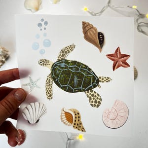 Peut inclure: Impression d'art carrée représentant une tortue de mer avec une carapace verte et marron, entourée de coquillages, d'une étoile de mer et de bulles. L'impression a un fond blanc et est tenue par une main. L'impression mesure environ 20 cm sur 20 cm.