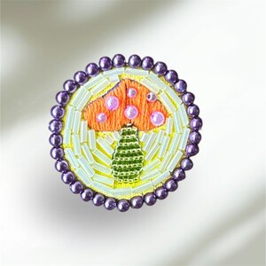 May include: A purple beaded brooch with a yellow background and an embroidered orange mushroom with pink dots.