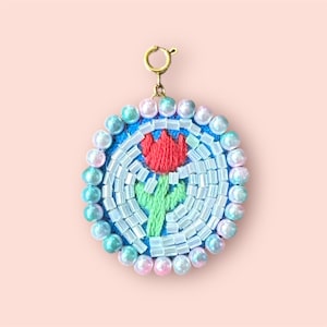 May include: A round, blue and white beaded charm with a red embroidered flower. The charm is attached to a gold chain.