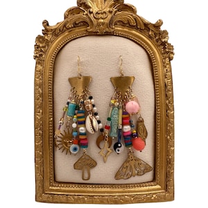 May include: A pair of gold-tone dangle earrings featuring a variety of colourful beads, charms, and pendants. The earrings are displayed on a gold-coloured stand.