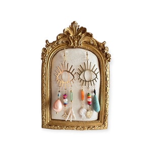 May include: A pair of gold eye-shaped earrings with colourful beads and charms hanging from them. The earrings are displayed on a gold ornate stand.
