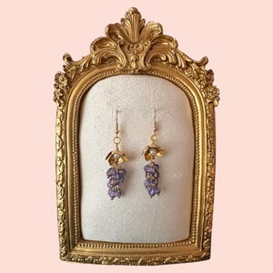 May include: A pair of gold earrings with purple flower-shaped charms and pearl accents. The earrings are displayed on a gold ornate frame.