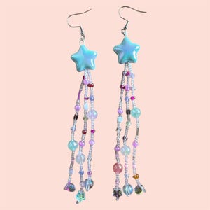May include: A pair of dangle earrings featuring a light blue iridescent star charm and a beaded fringe. The fringe is made of clear, pink, green, and purple beads.