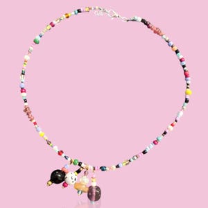 May include: A vibrant beaded necklace featuring a mix of small beads in pink, yellow, green, blue, and black. The necklace has charms, including a black bead, a white bead with black spots, and a purple bead. The necklace is set against a pink backdrop.