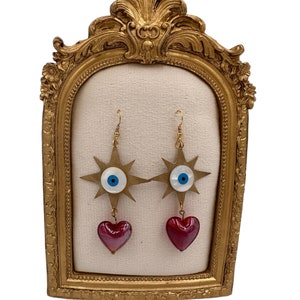 May include: Gold star-shaped earrings with a blue evil eye charm and a red glass heart dangle. The earrings are displayed on a gold-framed stand.