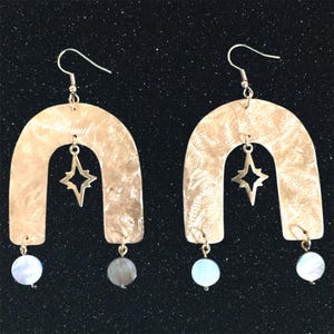 May include: A pair of gold-toned earrings with a hammered metal arch shape and a star charm hanging from the centre. Each earring has a white pearl-like bead dangling from the bottom.