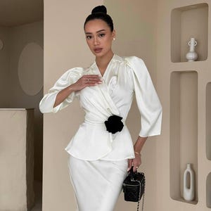 May include: A white satin blouse with a black flower accent at the waist. The blouse has a wrap style and long sleeves with puffed shoulders. The model is wearing a matching white skirt.