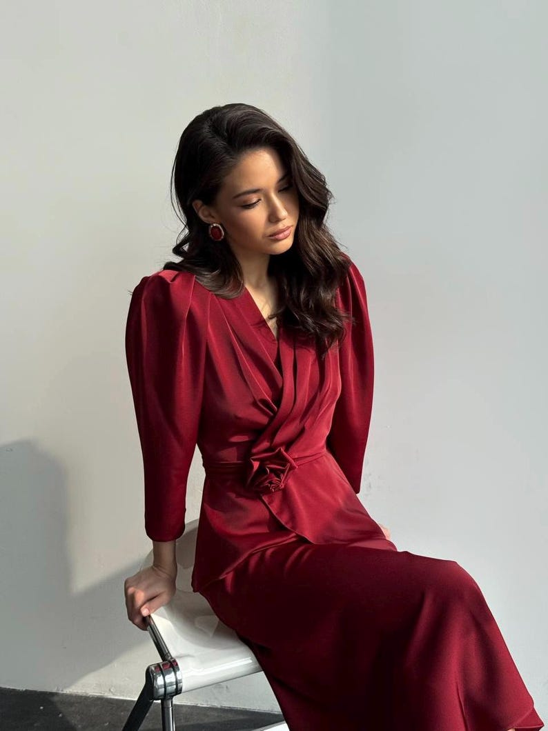 May include: A woman wearing a long-sleeved, burgundy dress with a cinched waist and a large flower detail. The dress has a V-neckline and a slightly puffed sleeve.