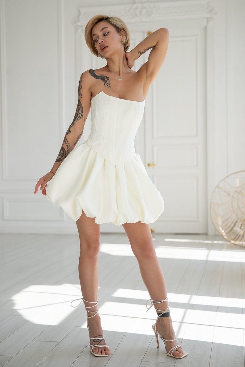 May include: A strapless white dress with a fitted bodice and a bubble skirt. The dress is paired with white strappy high-heeled sandals. The dress has a structured, corset-like bodice and a voluminous skirt. The sandals have thin straps that wrap around the ankles.