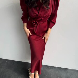 May include: A long, burgundy satin dress with a wrap-style bodice and a large flower accent at the waist. The dress has long sleeves with puffed shoulders and a fitted skirt.