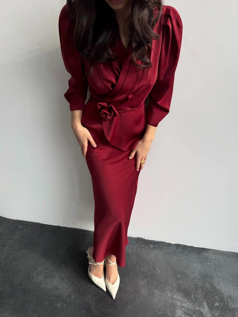 May include: A long, burgundy satin dress with a wrap-style bodice and a large flower accent at the waist. The dress has long sleeves with puffed shoulders and a fitted skirt.