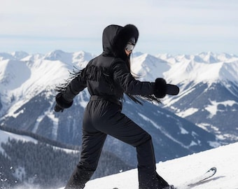 May include: A person skiing downhill in a black snowsuit with fringe detail on the sleeves and hood. The snowsuit has a belt and is paired with black gloves and goggles. The background features snow-covered mountains and a blue sky.
