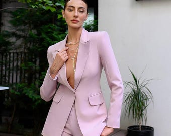 Könnte beinhalten: Ein hellrosa Anzug, bestehend aus einem Blazer und einer passenden Hose. Der Blazer hat ein fallendes Revers und zwei Vordertaschen. Eine goldene Halskette ist sichtbar. Der Anzug wird im Freien getragen, mit Grün im Hintergrund.