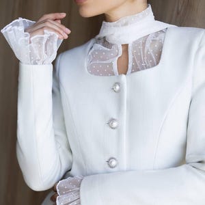 May include: A white blazer with a square neckline and pearl-like buttons. The blazer features sheer, dotted fabric at the neckline and cuffs, creating a layered effect. The sleeves are long and fitted.