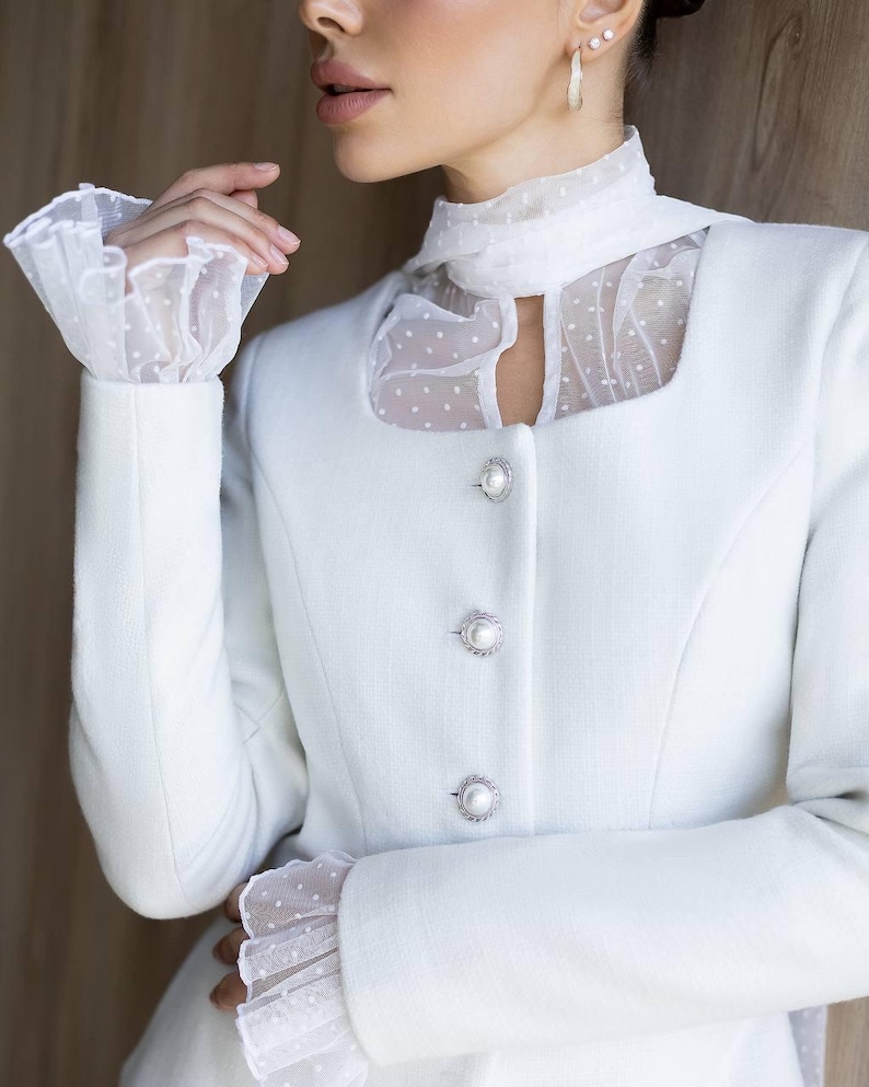 May include: A white blazer with a square neckline and pearl-like buttons. The blazer features sheer, dotted fabric at the neckline and cuffs, creating a layered effect. The sleeves are long and fitted.