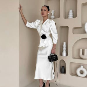 May include: A woman wearing a white satin dress with a black flower accent at the waist. She is holding a black chain bag and wearing black high heels.