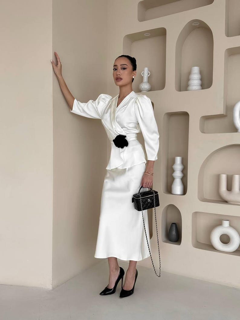 May include: A woman wearing a white satin dress with a black flower accent at the waist. She is holding a black chain bag and wearing black high heels.