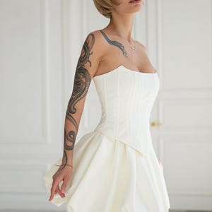 May include: A white strapless corset-style dress with a sweetheart neckline and a short, bubble skirt. The dress is fitted at the bodice and flares out at the waist. The woman has a snake tattoo on her arm and a short blonde hairstyle.