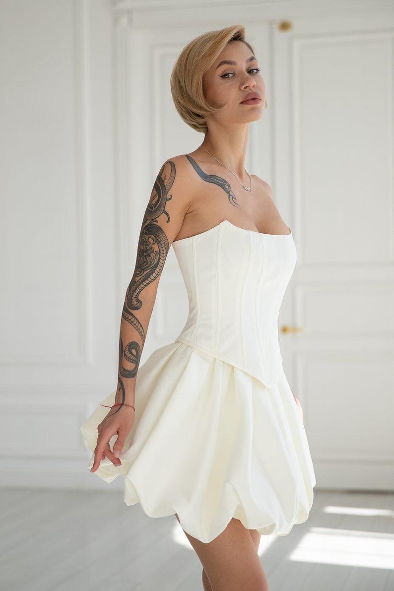 May include: A white strapless corset-style dress with a sweetheart neckline and a short, bubble skirt. The dress is fitted at the bodice and flares out at the waist. The woman has a snake tattoo on her arm and a short blonde hairstyle.
