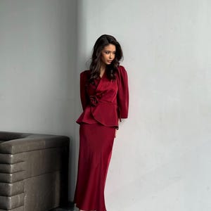May include: A woman wearing a long, burgundy satin skirt and matching top with a bow detail. She is wearing white heels.