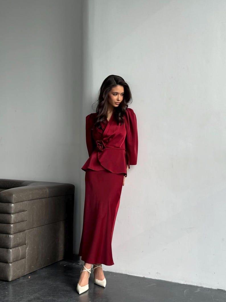 May include: A woman wearing a long, burgundy satin skirt and matching top with a bow detail. She is wearing white heels.