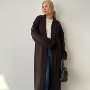 Könnte beinhalten: Ein langer, dunkelbrauner Strickcardigan mit offener Vorderseite und gerippten Bündchen, getragen über einem weißen Ripp-Oberteil und einer blauen Jeans. Das Outfit wird durch eine kleine schwarze Stepptasche mit goldener Kette ergänzt.