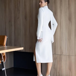May include: A white skirt suit with a high-neck jacket and a side slit skirt. The jacket has long sleeves and a flowing back detail. The outfit is paired with light-colored high heels. The background is a wooden wall.