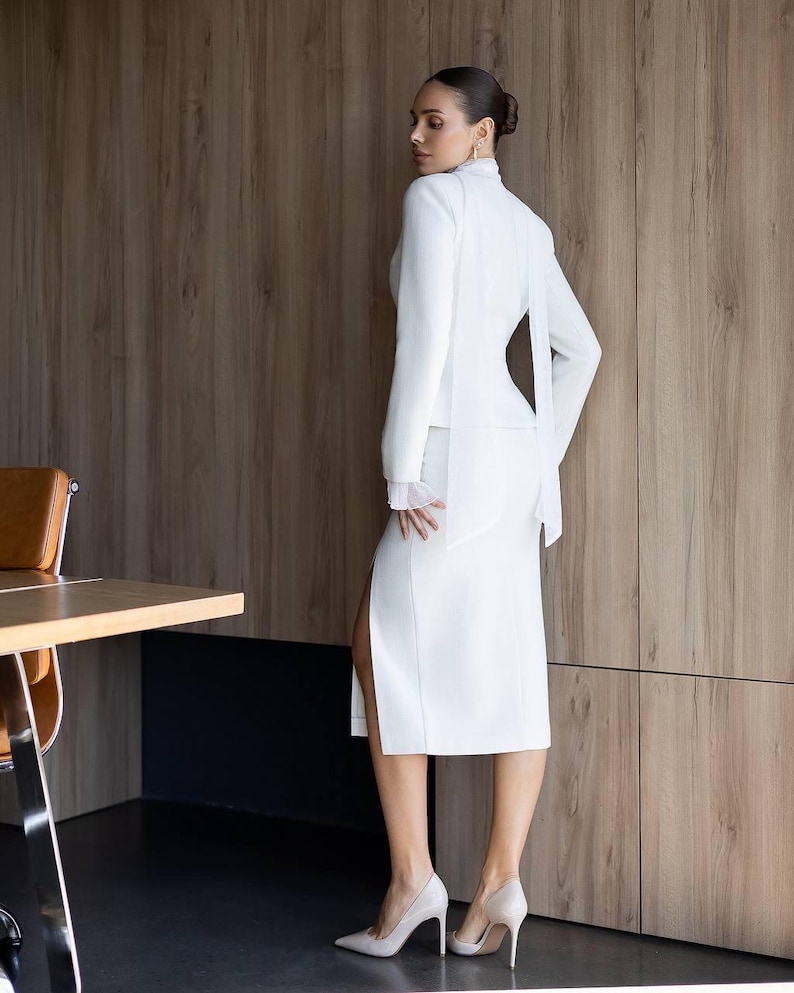 May include: A white skirt suit with a high-neck jacket and a side slit skirt. The jacket has long sleeves and a flowing back detail. The outfit is paired with light-colored high heels. The background is a wooden wall.