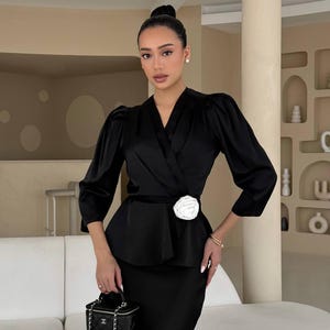 May include: A black satin blouse with a white flower accent at the waist. The blouse has a wrap style and long sleeves. The model is wearing a black skirt and carrying a small black handbag.