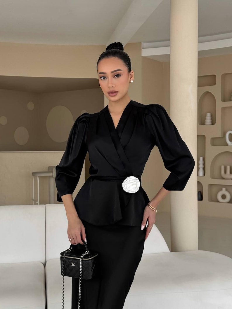 May include: A black satin blouse with a white flower accent at the waist. The blouse has a wrap style and long sleeves. The model is wearing a black skirt and carrying a small black handbag.