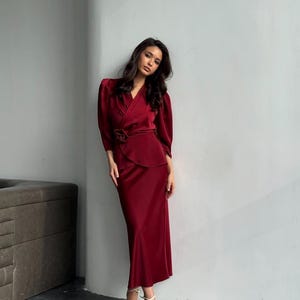 May include: A woman wearing a long, burgundy satin dress with a wrap-style bodice and a matching skirt. The dress has a cinched waist and a flowing skirt. She is wearing white heels.