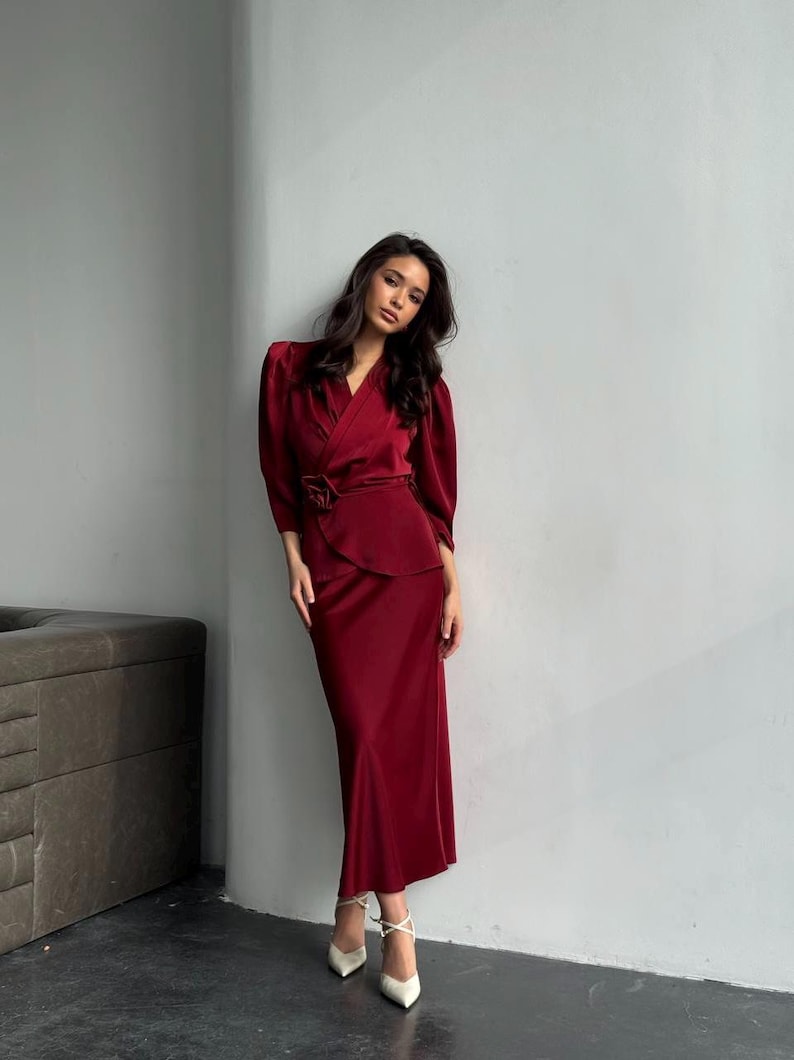 May include: A woman wearing a long, burgundy satin dress with a wrap-style bodice and a matching skirt. The dress has a cinched waist and a flowing skirt. She is wearing white heels.