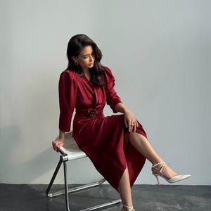 May include: A woman wearing a burgundy satin dress with a cinched waist and puff sleeves sits on a white metal chair. She is wearing white heels with ankle straps.