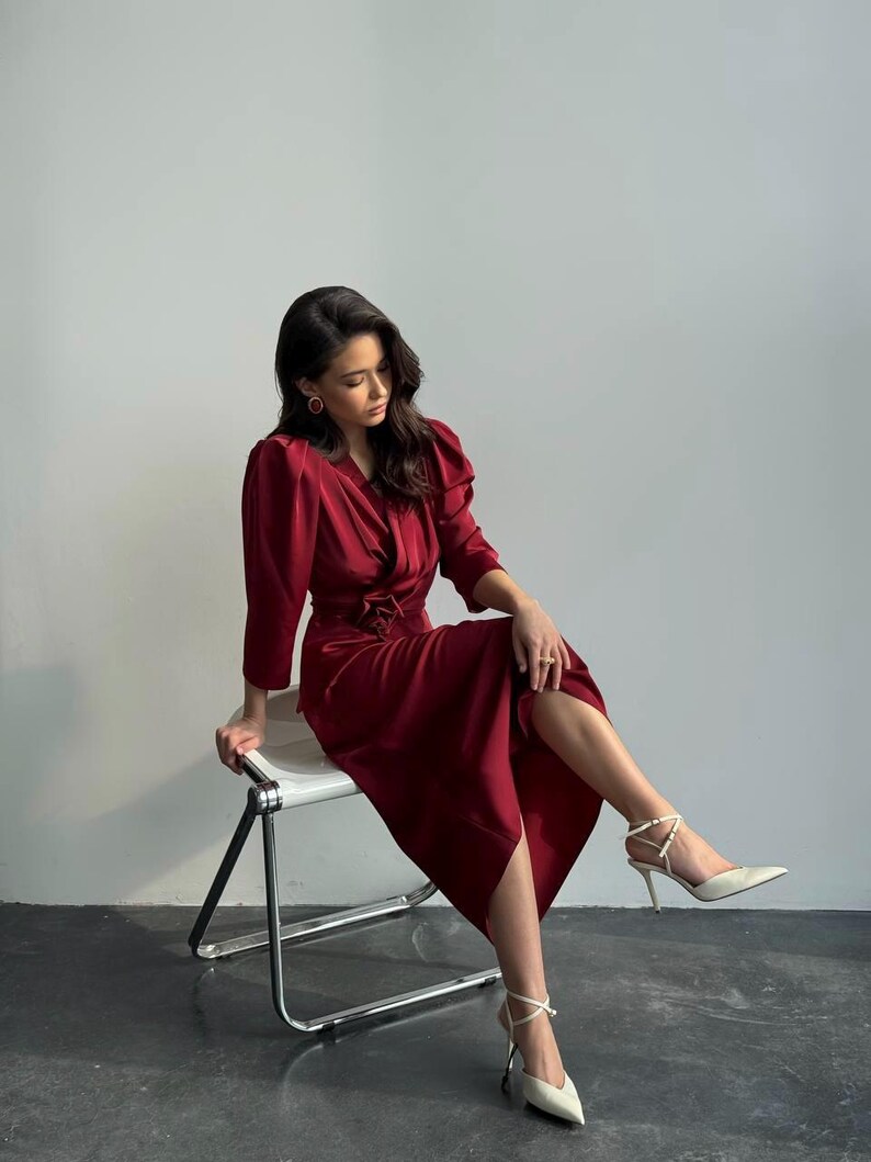 May include: A woman wearing a burgundy satin dress with a cinched waist and puff sleeves sits on a white metal chair. She is wearing white heels with ankle straps.