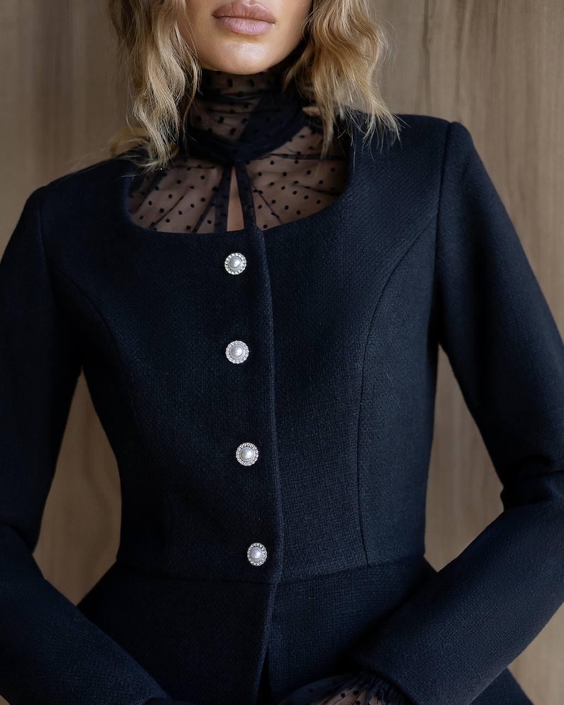 May include: A black blazer with a square neckline and three pearl-like buttons. The blazer features a sheer, polka-dot panel at the neckline. The sleeves are long and the fabric has a textured appearance.
