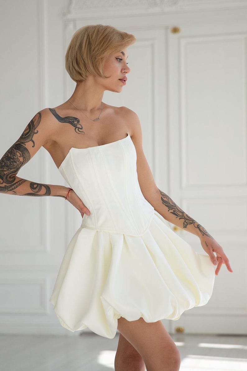 May include: A strapless, ivory-colored corset dress. The bodice is fitted with vertical seams, and the skirt is a voluminous bubble shape. The dress is made of a smooth, opaque fabric. The model has short blonde hair and tattoos on their arms.