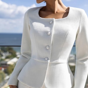 May include: A white, tailored suit featuring a square neckline jacket with pearl-like buttons. The jacket has a peplum design, and the suit appears to be made of a textured fabric. The background shows a view of the ocean and sky.