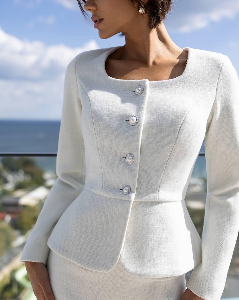 May include: A white, tailored suit featuring a square neckline jacket with pearl-like buttons. The jacket has a peplum design, and the suit appears to be made of a textured fabric. The background shows a view of the ocean and sky.
