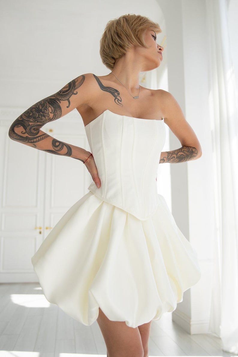 May include: A strapless white corset dress with a bubble skirt. The corset bodice has vertical seams. The skirt is voluminous and gathered at the waist, creating a rounded shape. The dress is made of a smooth, opaque fabric.