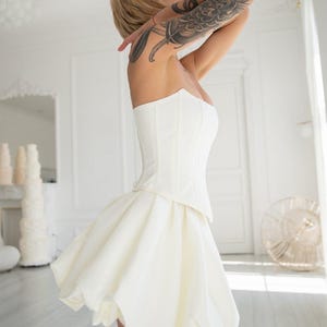 May include: A strapless white corset top paired with a short, flared white skirt. The skirt has a bubble hem. The woman has short blonde hair and a large tattoo on her arm. The background is a bright white room with a window.