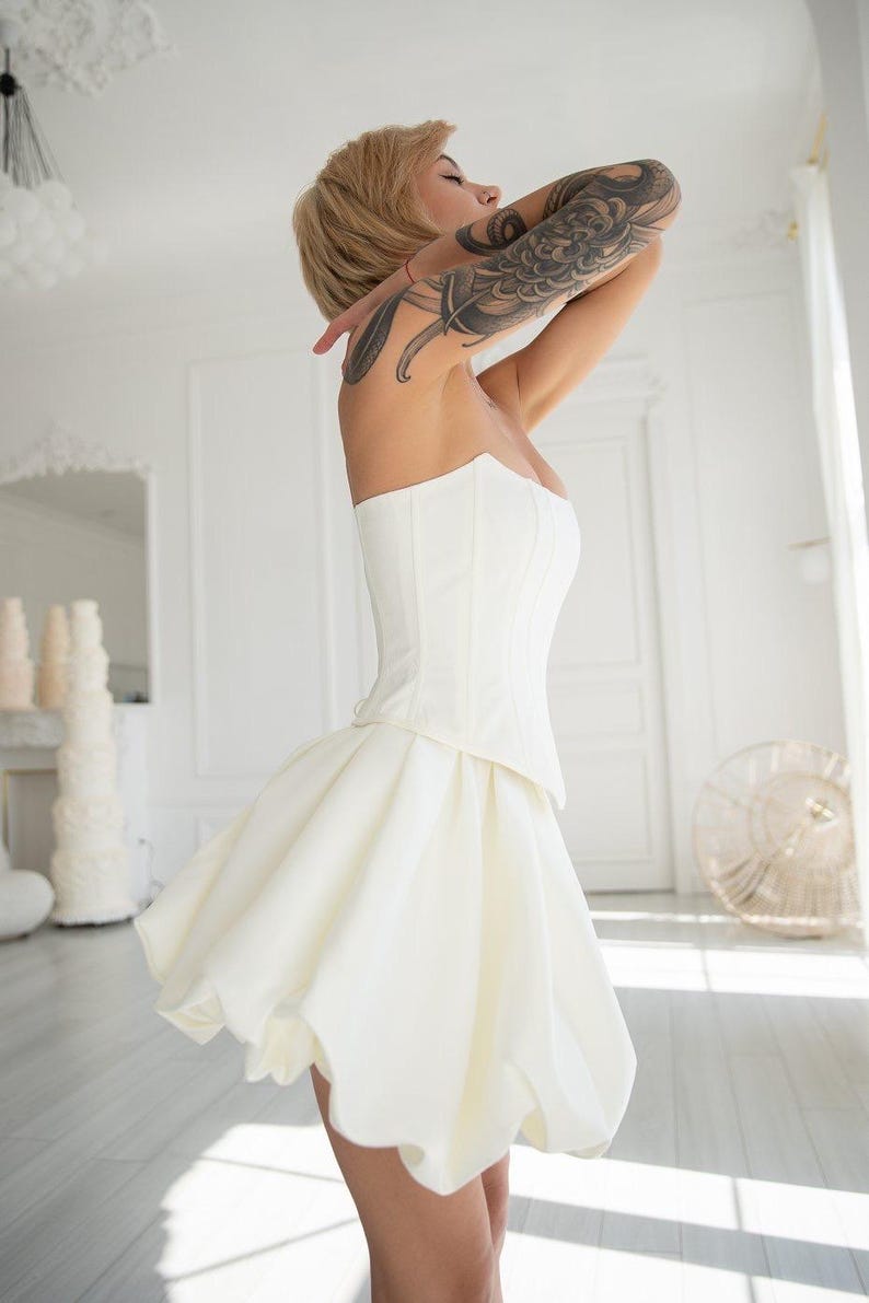 May include: A strapless white corset top paired with a short, flared white skirt. The skirt has a bubble hem. The woman has short blonde hair and a large tattoo on her arm. The background is a bright white room with a window.
