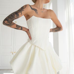 May include: A strapless white corset-style dress with a bubble skirt. The bodice is fitted with vertical seams, and the skirt is voluminous, gathered at the waist, and rounded at the hem. The dress is made of a smooth, opaque fabric.