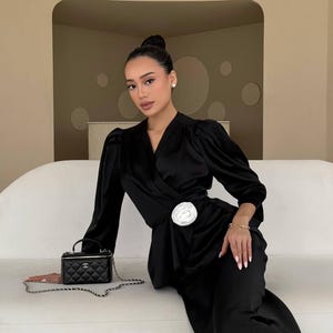 May include: A woman wearing a black satin wrap dress with a white flower brooch. She is sitting on a white couch and holding a black Chanel handbag with a silver chain.