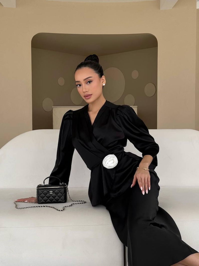 May include: A woman wearing a black satin wrap dress with a white flower brooch. She is sitting on a white couch and holding a black Chanel handbag with a silver chain.