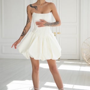 May include: A strapless white mini dress with a corset-style bodice and a voluminous bubble skirt. The dress is paired with white strappy heeled sandals. The model has short blonde hair and tattoos on both arms. The background is a white room with a door.