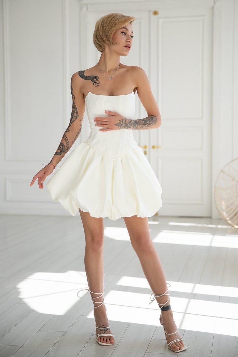 May include: A strapless white mini dress with a corset-style bodice and a voluminous bubble skirt. The dress is paired with white strappy heeled sandals. The model has short blonde hair and tattoos on both arms. The background is a white room with a door.