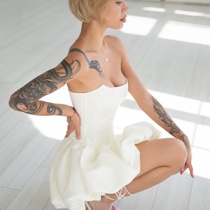 May include: A strapless white corset-style dress with a ruffled skirt. The dress is paired with white, strappy high-heeled sandals. The person has short, blonde hair and tattoos on their arms.