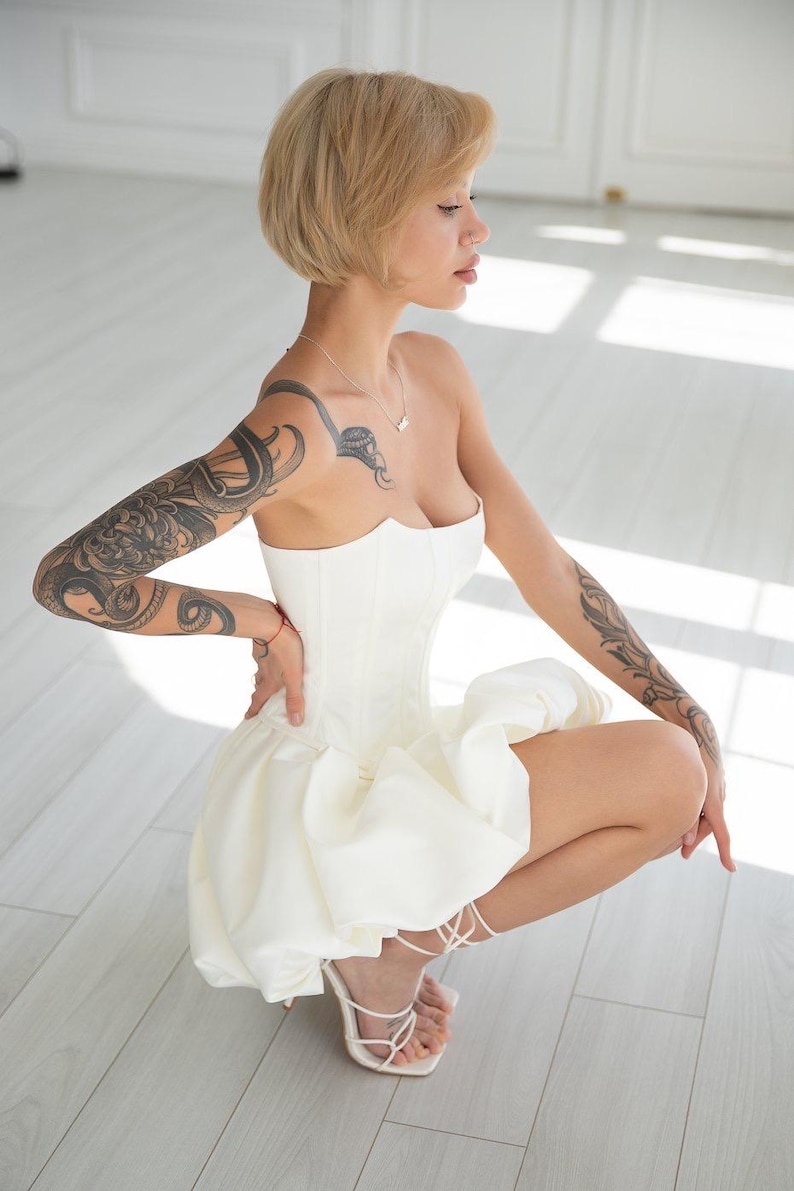 May include: A strapless white corset-style dress with a ruffled skirt. The dress is paired with white, strappy high-heeled sandals. The person has short, blonde hair and tattoos on their arms.