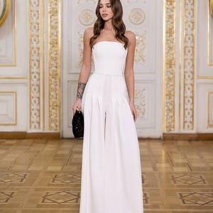 May include: A strapless white jumpsuit with wide-leg pants. The outfit features a fitted bodice and a thin belt at the waist. The person is holding a black sequined clutch. The background is a room with ornate gold trim and a patterned wooden floor.